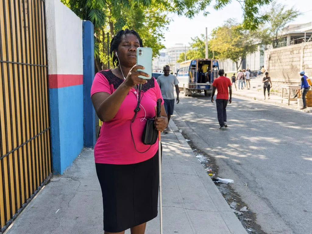 Una mujer con bastón blanco sostiene su teléfono en vertical frente al pecho, con la cámara hacia la calle.